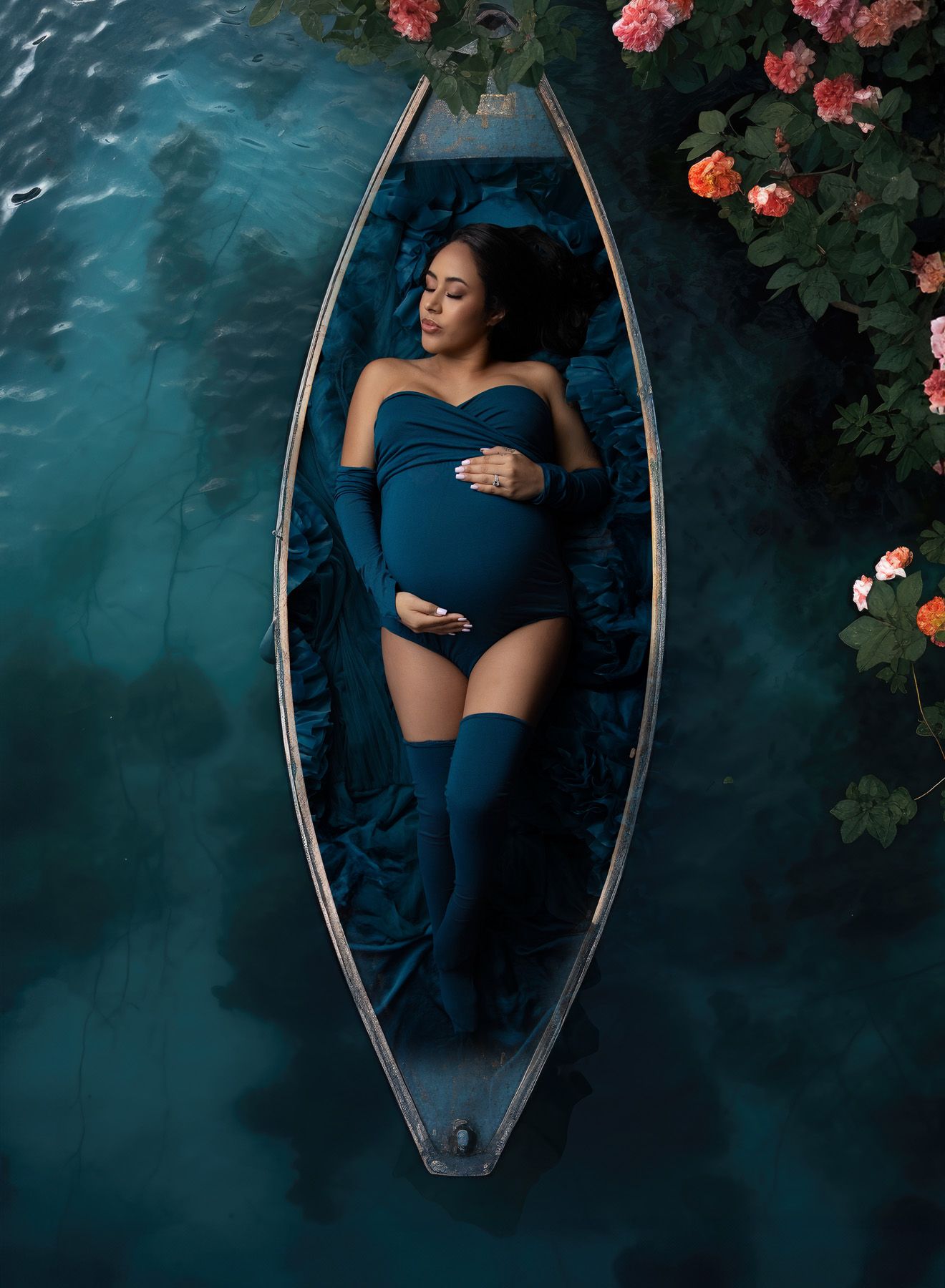 Maternity portrait of mom laying in a boat in teal bodysuit and stockings  the boat is in a teal sea surrounded by pink flowers.