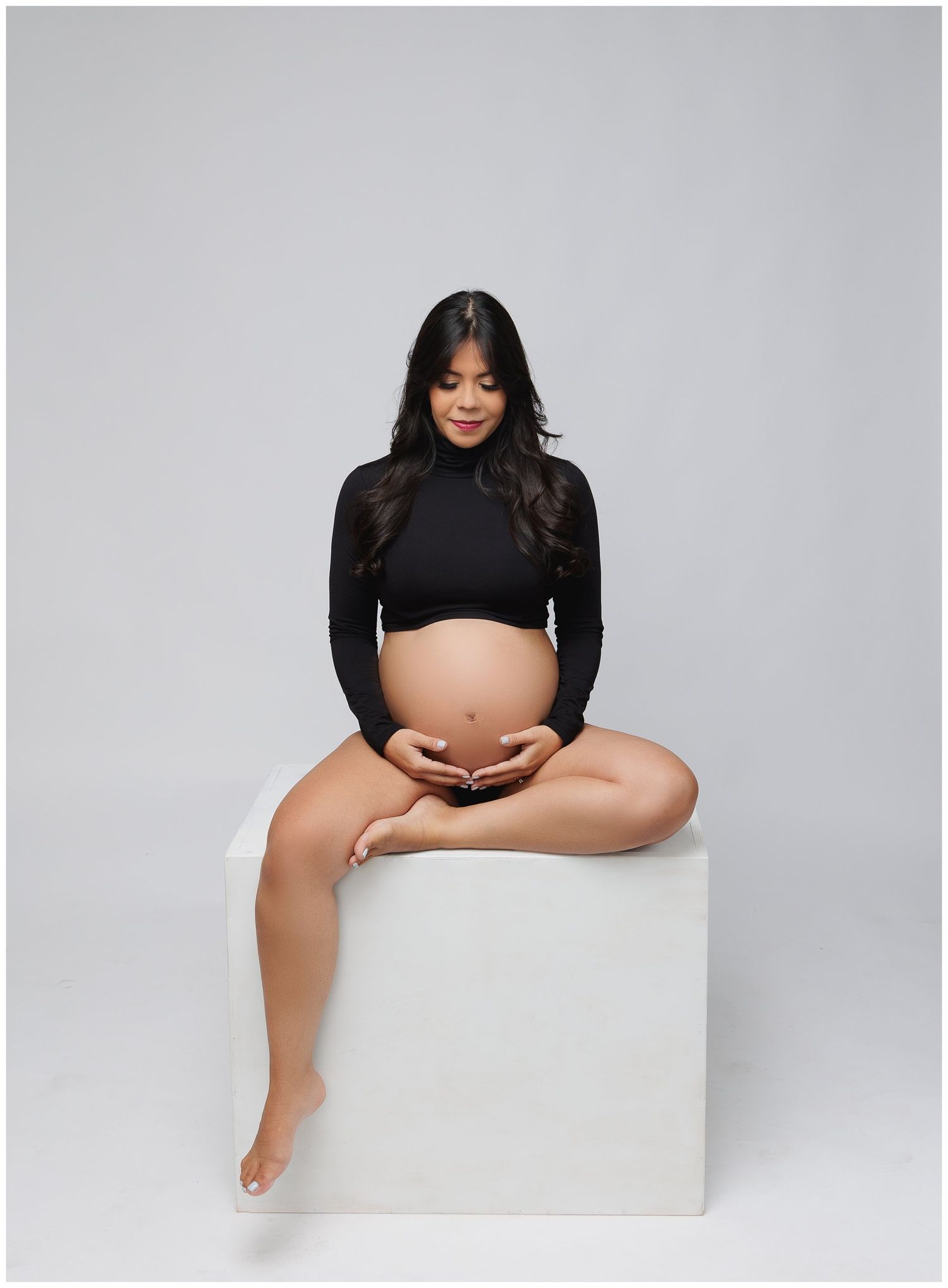Modern maternity portrait of mom seated cross legged on top of white posing cube wearing a black crop top with belly exposed.
