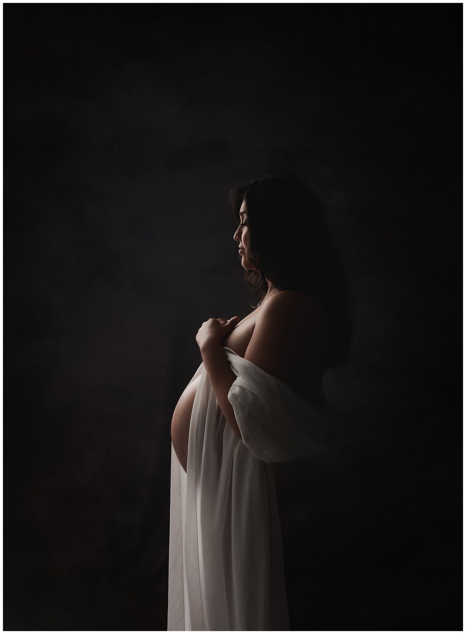 Austin maternity photographer studio portrait of pregnant mother in flowing sheer fabric creating dramatic silhouette against dark backgroun