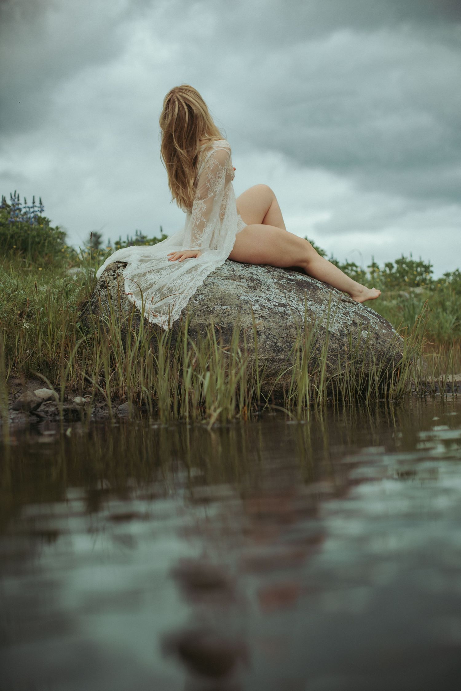 Woman in white lace dress sitting on a boulder by a calm lake under cloudy skies.
