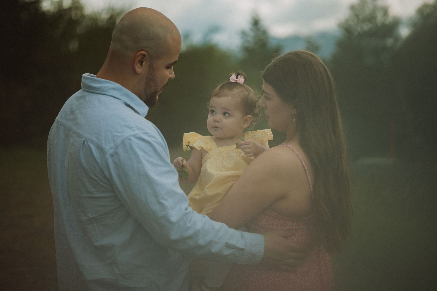 Parents lovingly gaze at their baby daughter dressed in yellow outdoors in soft light, in Alaska.