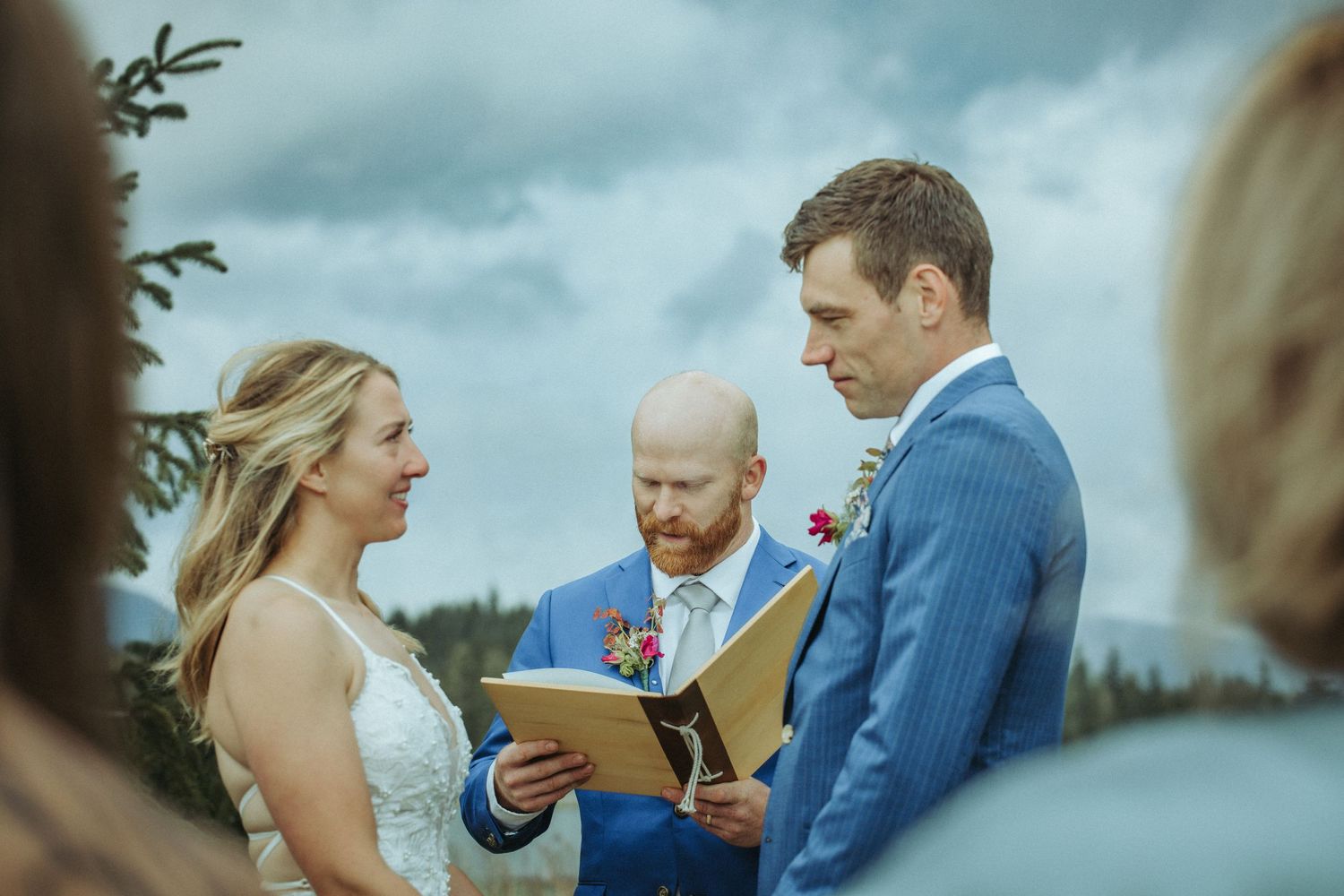 An intimate outdoor wedding ceremony takes place against a backdrop of evergreen trees and moody clouds in Alaska.