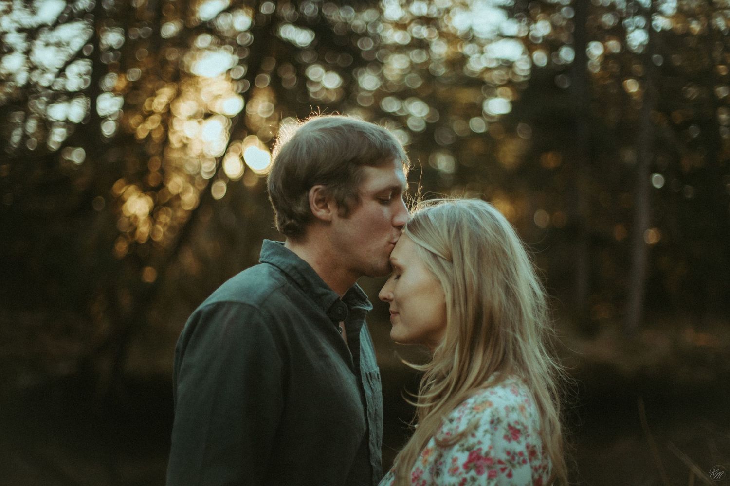 A couple shares an intimate moment at sunset in a wooded area with golden light filtering through the trees in Southeast Alaska