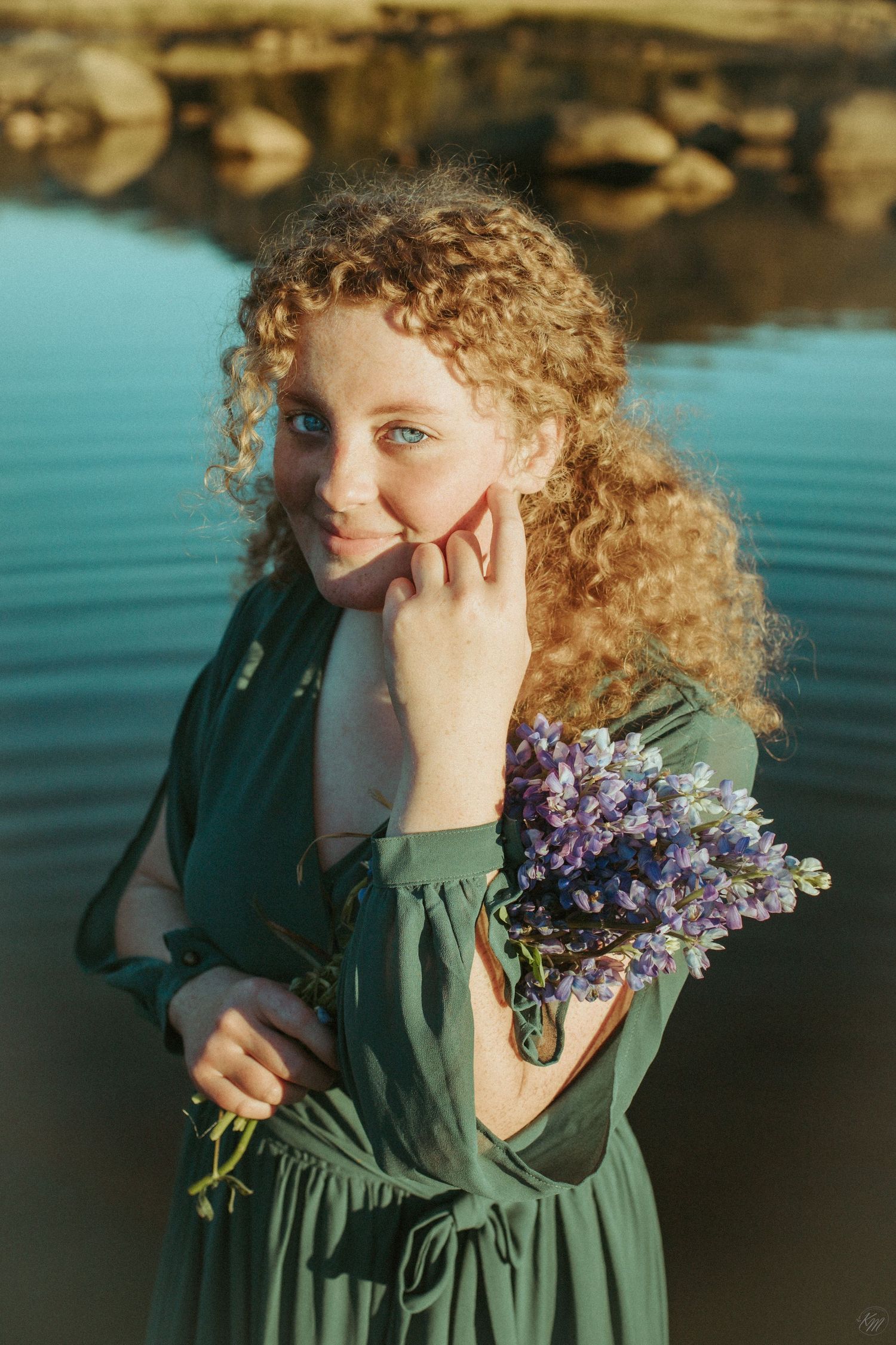 Ethereal portrait in green dress holding purple flowers by calm lake waters.