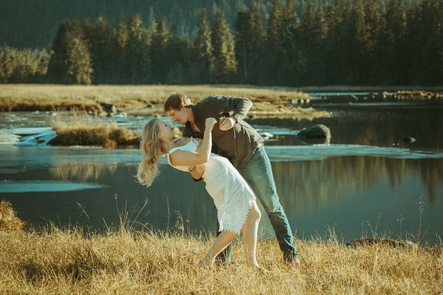 A couple plays together in a golden field next to a serene lake with mountains and forest in the background in Southeast Alaska.