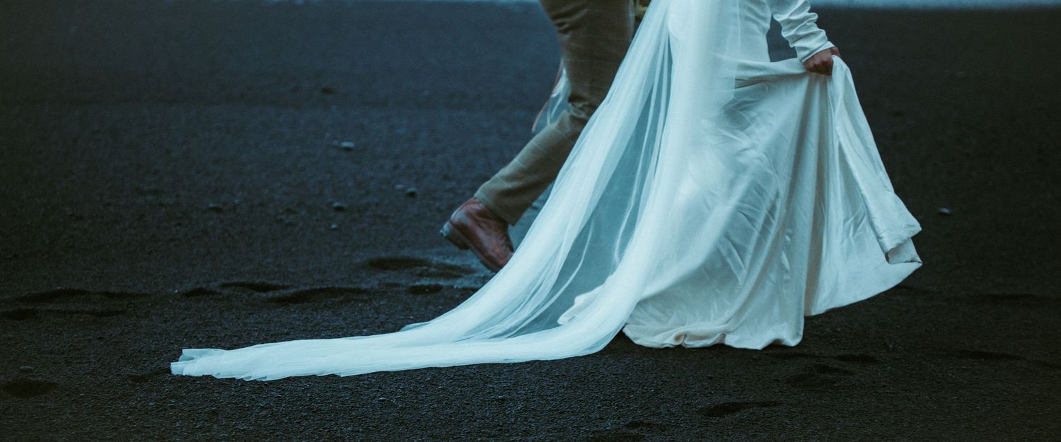 Bride in flowing white wedding gown walking on black sand beach leaving footprints behind.