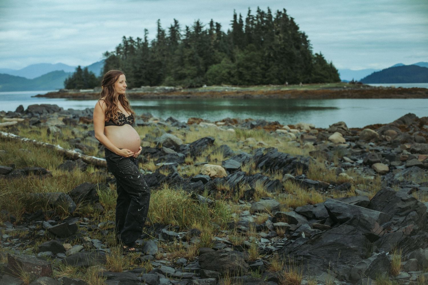 Pregnant woman cradling her belly on rocky Alaskan shoreline with island and mountains behind her.