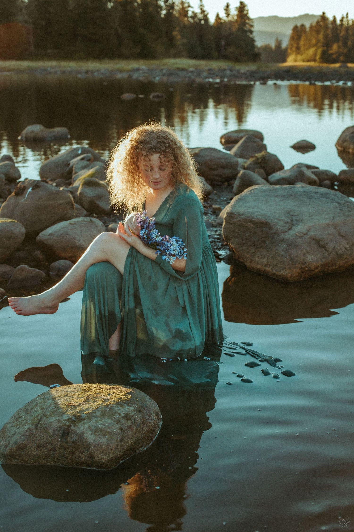 A person in a teal dress sits on rocks by a serene lake at sunset holding blue flowers.