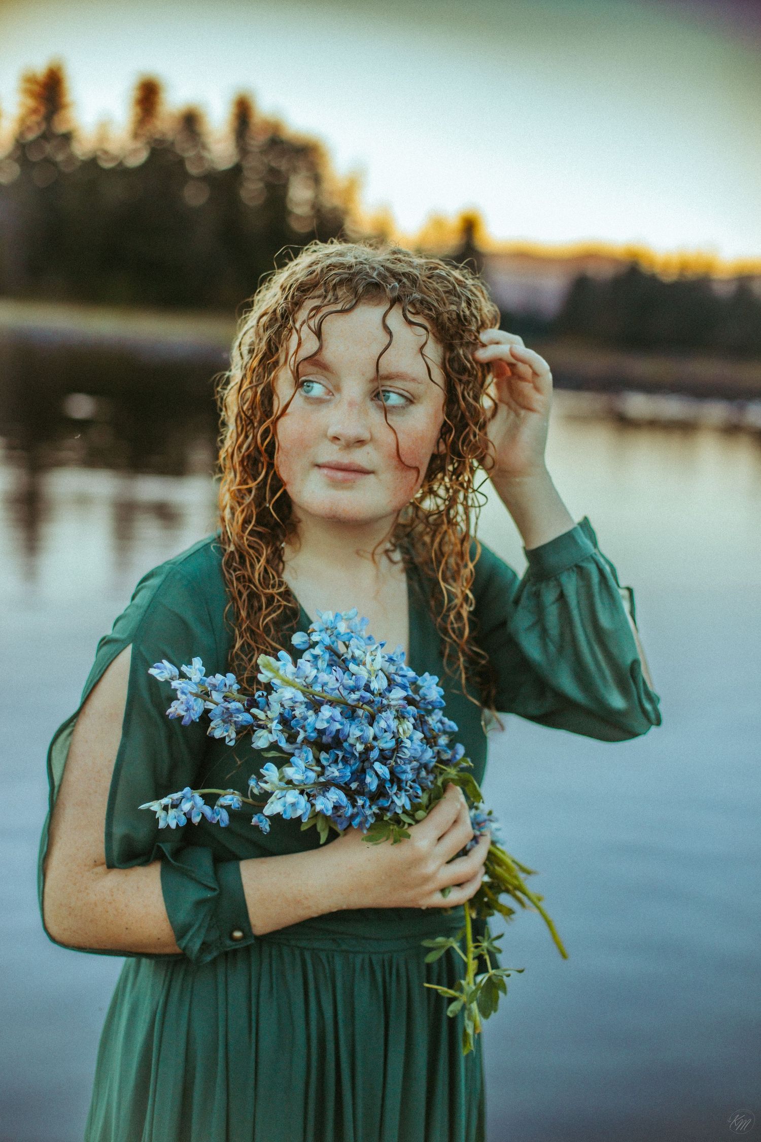 A green-dressed figure holds a bouquet of blue flowers by a lake at sunset.