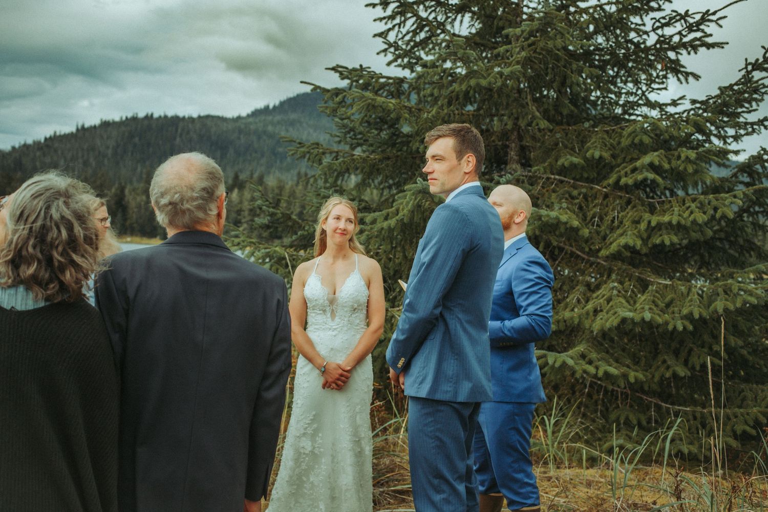 Outdoor wedding ceremony unfolds against evergreen trees and dramatic cloudy skies in Alaska.