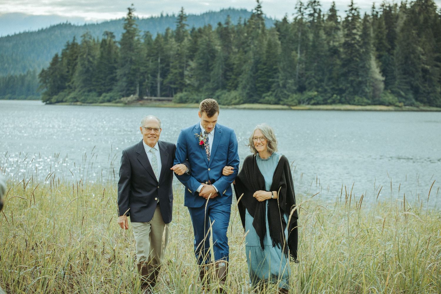 Three people walk together in tall grass by a serene lake surrounded by evergreen forest.