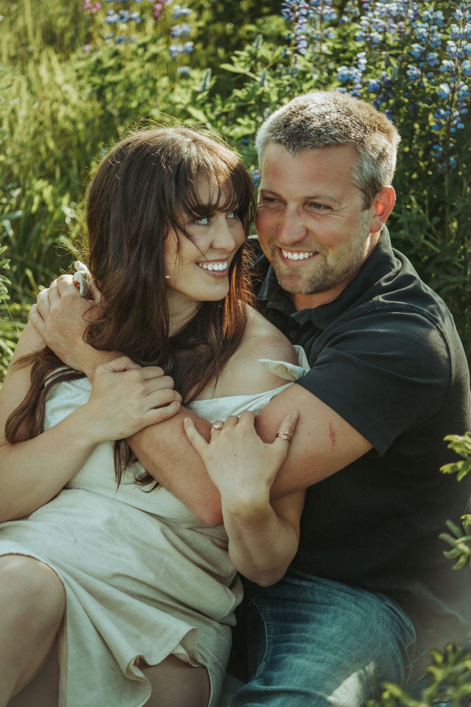 A romantic engaged couple embrace outdoors on a sunny day in Alaska, with blue flowers, lupine and greenery in the background.