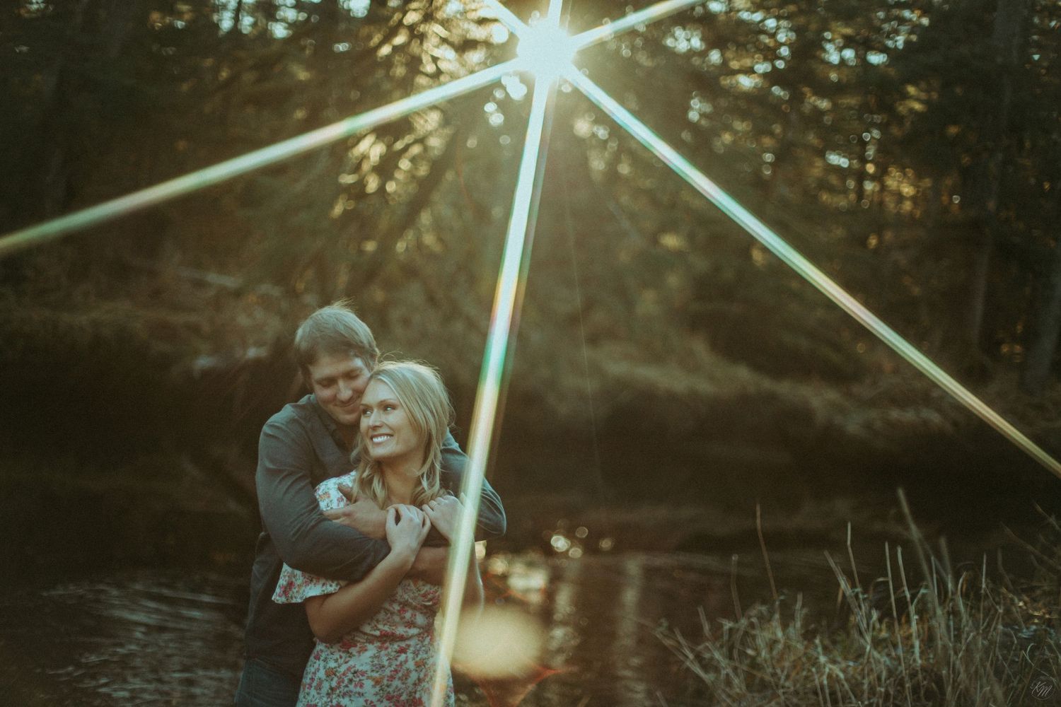 Romantic couple embraces by a lake at sunset in Southeast Alaska, with dramatic light rays filtering through trees during golden hour
