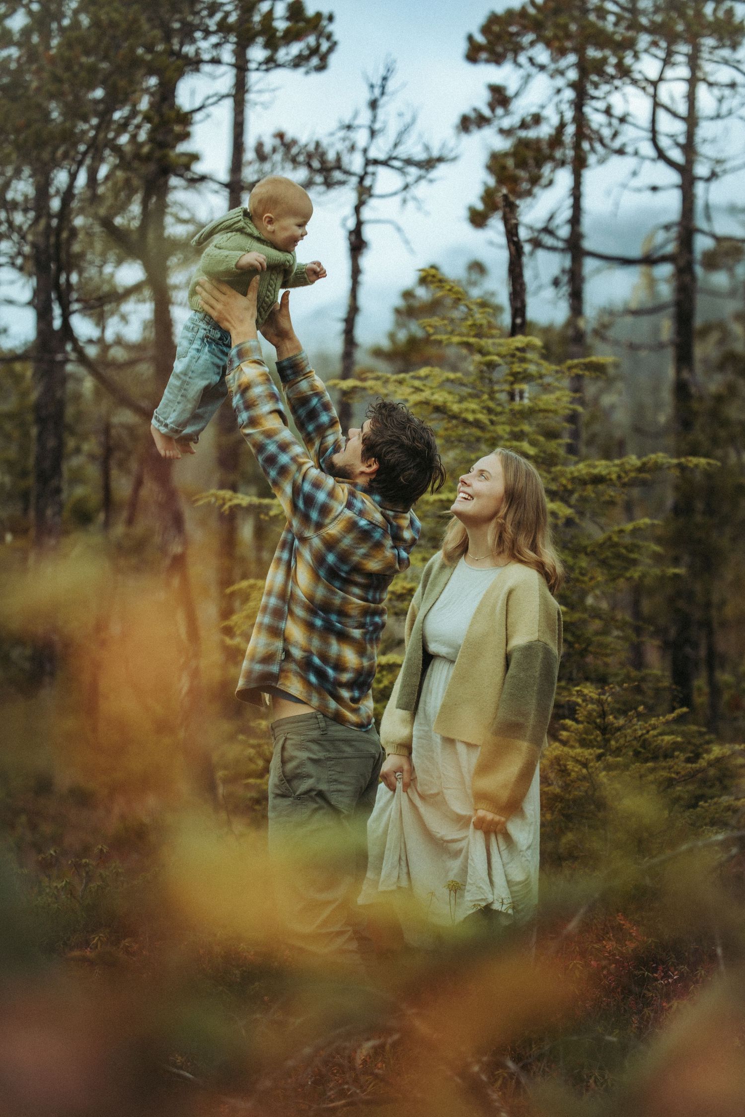 A joyful family in a forest, father lifting baby overhead while mother laughs, surrounded by autumn foliage and pine trees.