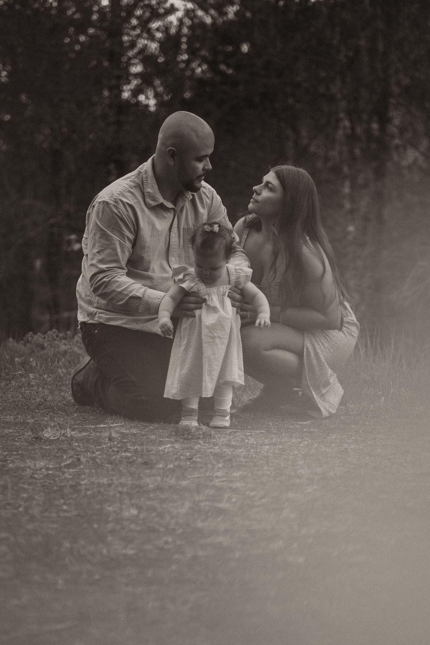 Sepia-toned family portrait of parents crouching with toddler girl in white dress outdoors in nature.