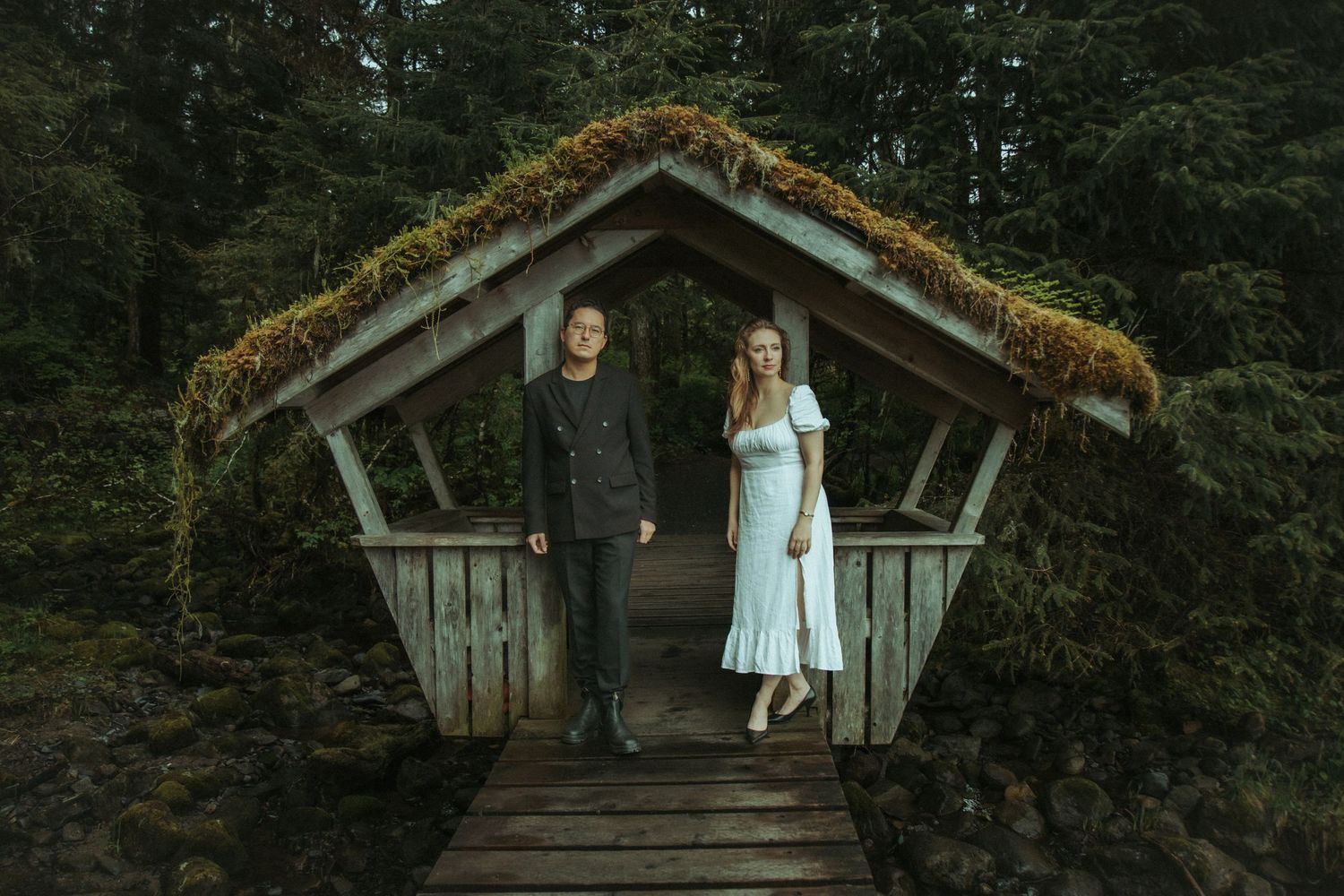 Couple stands together on a moss-covered wooden bridge in Southeast Alaska surrounded by lush evergreen forest.