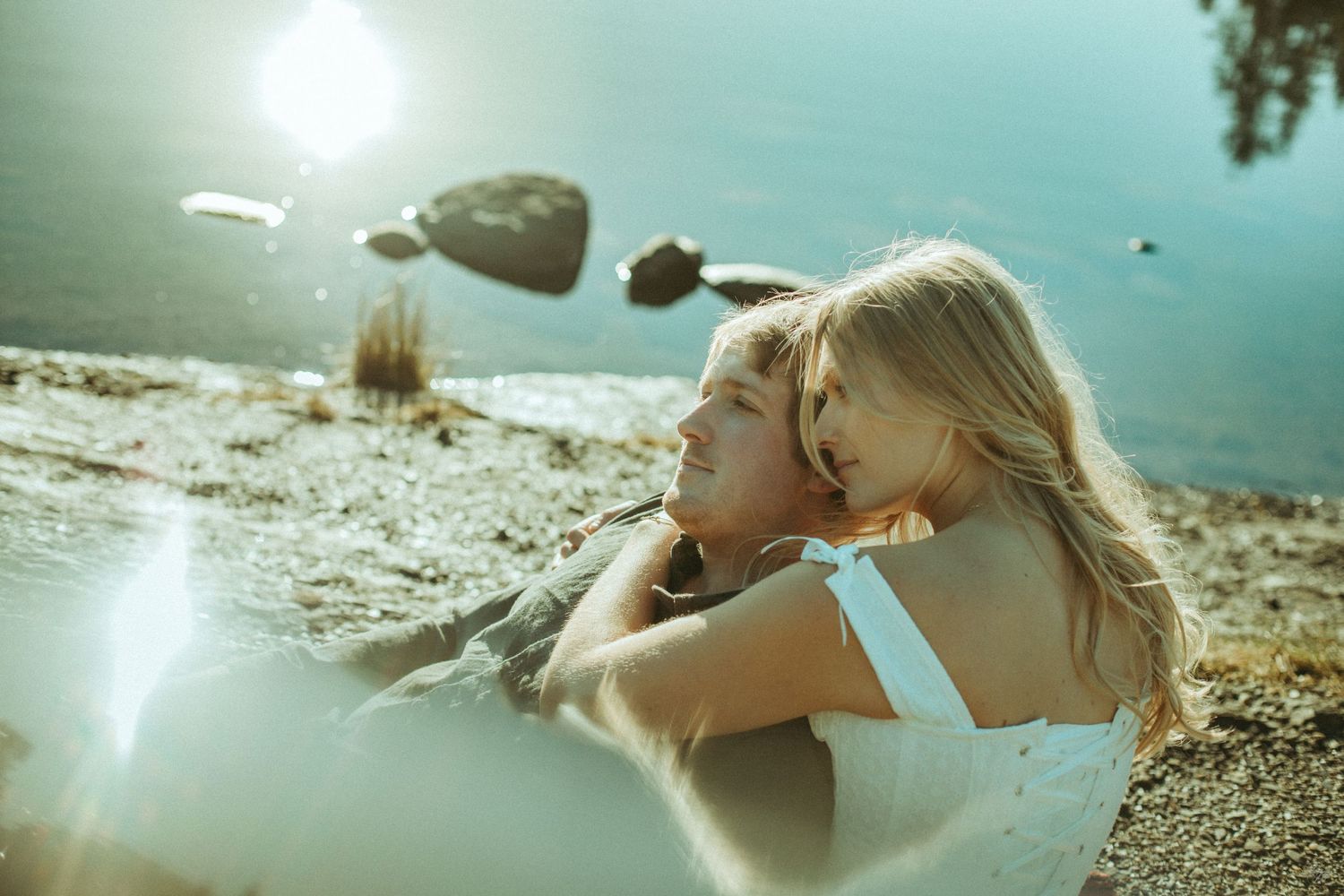 Two people share an intimate moment by a lake while sun flares create a dreamy atmosphere at golden hour in Southeast Alaska.