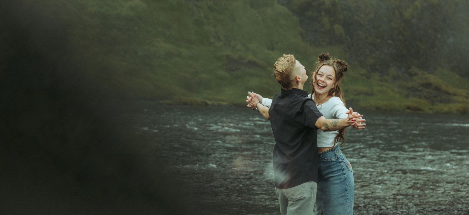 Joyful couple embracing and laughing together near a river in a lush green outdoor setting.