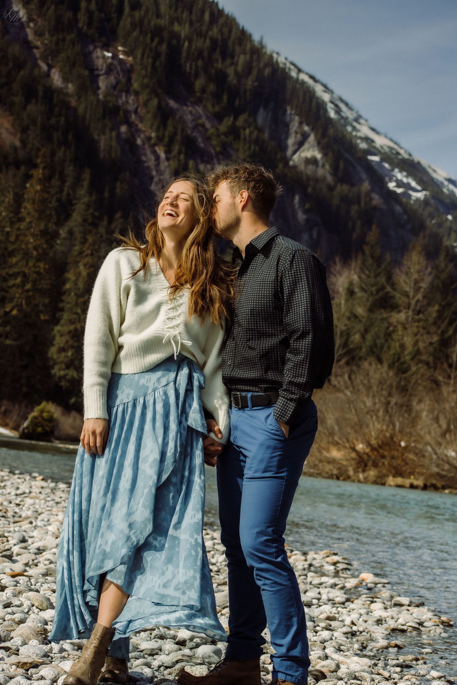Happy couple sharing intimate moment on rocky riverbank with mountain landscape behind them.