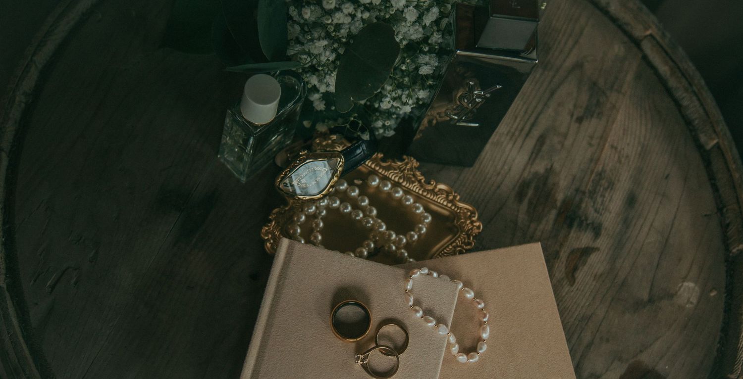 Elegant wedding flat lay with pearl necklace, rings, mirror, and floral arrangement on dark surface.