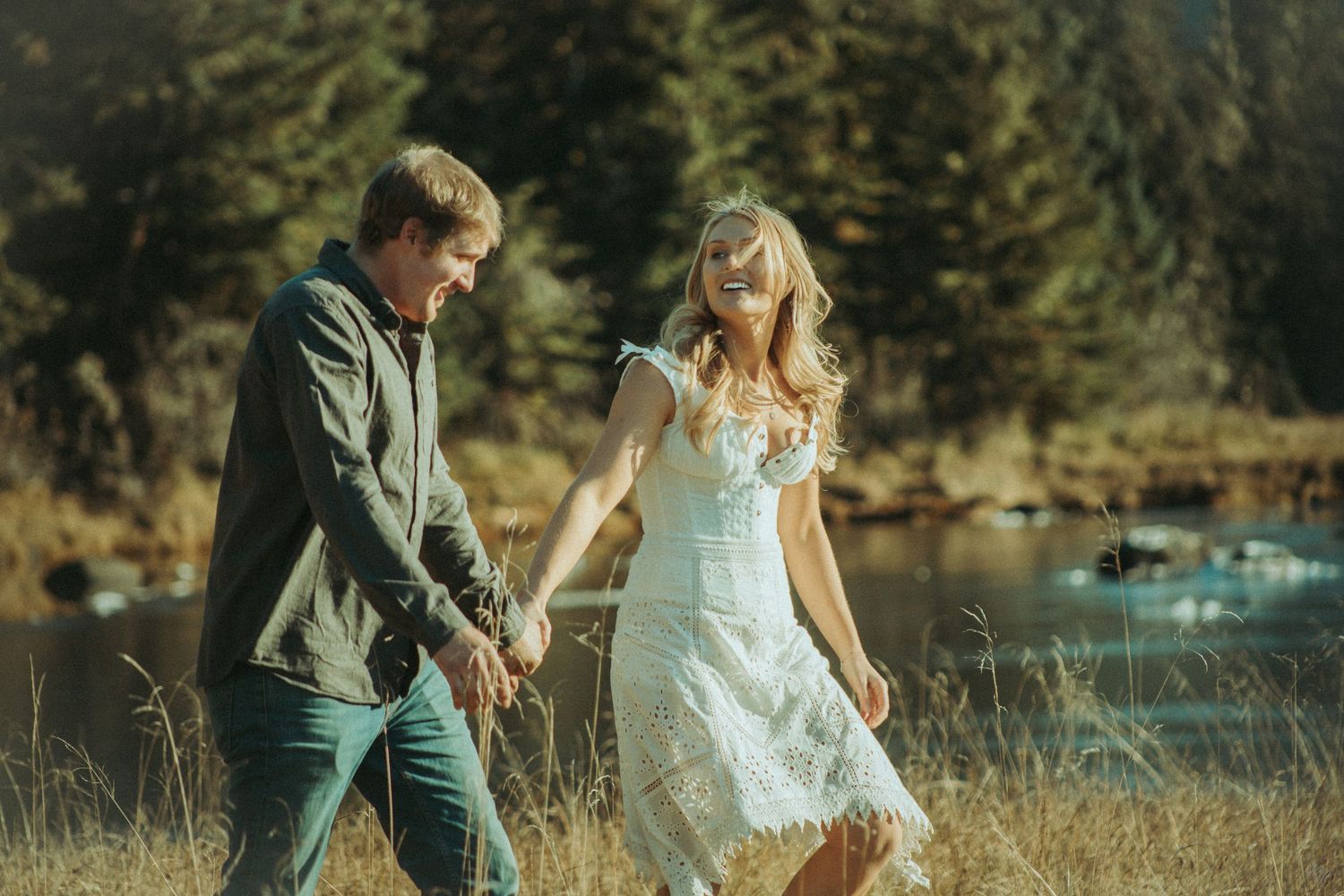 A couple walks hand in hand through a sunlit meadow near a lake with evergreen trees in the background in Southeast Alaska