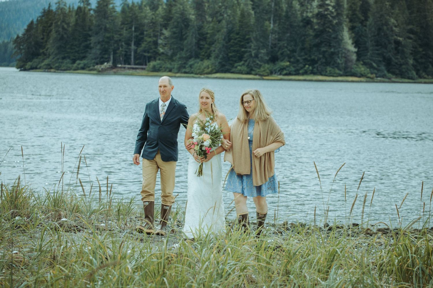 Wedding party stands together on lakeside shore with mountains and forest in background in Alaska.
