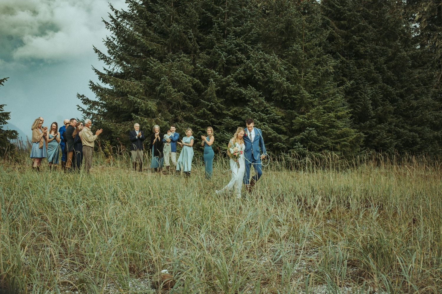 Wedding party and guests walk along a grassy beach path near evergreen trees in Alaska.