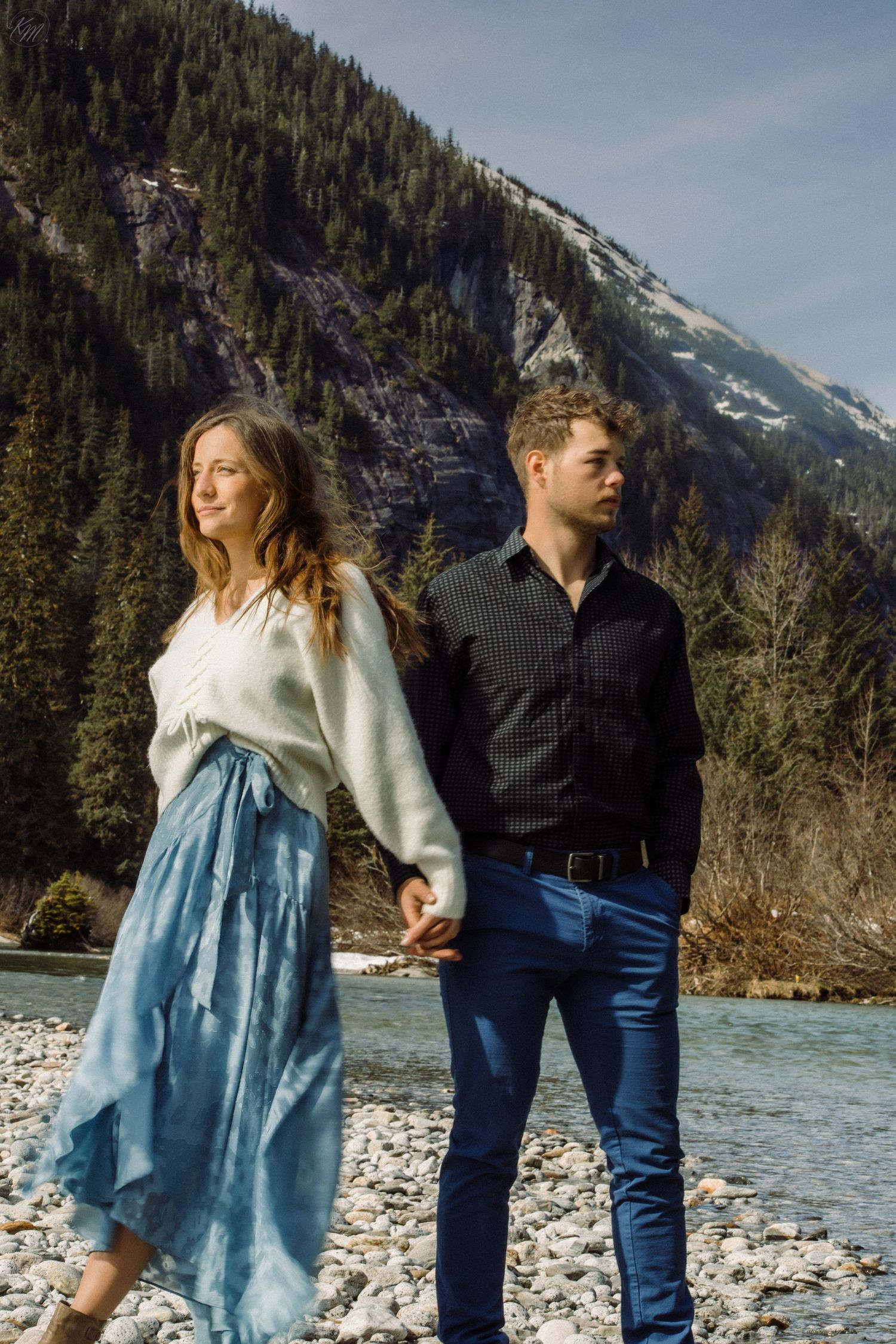 Stylish couple in casual attire walking along rocky riverbank with mountain vista in background.