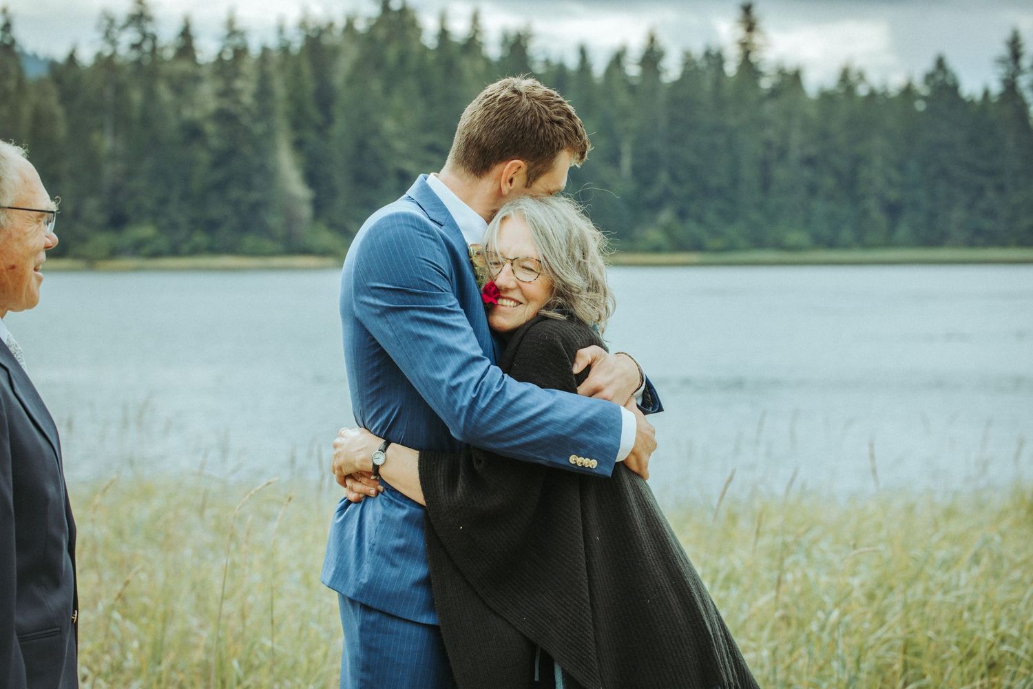 People embrace emotionally beside a scenic mountain lake as evergreen trees line the shore.