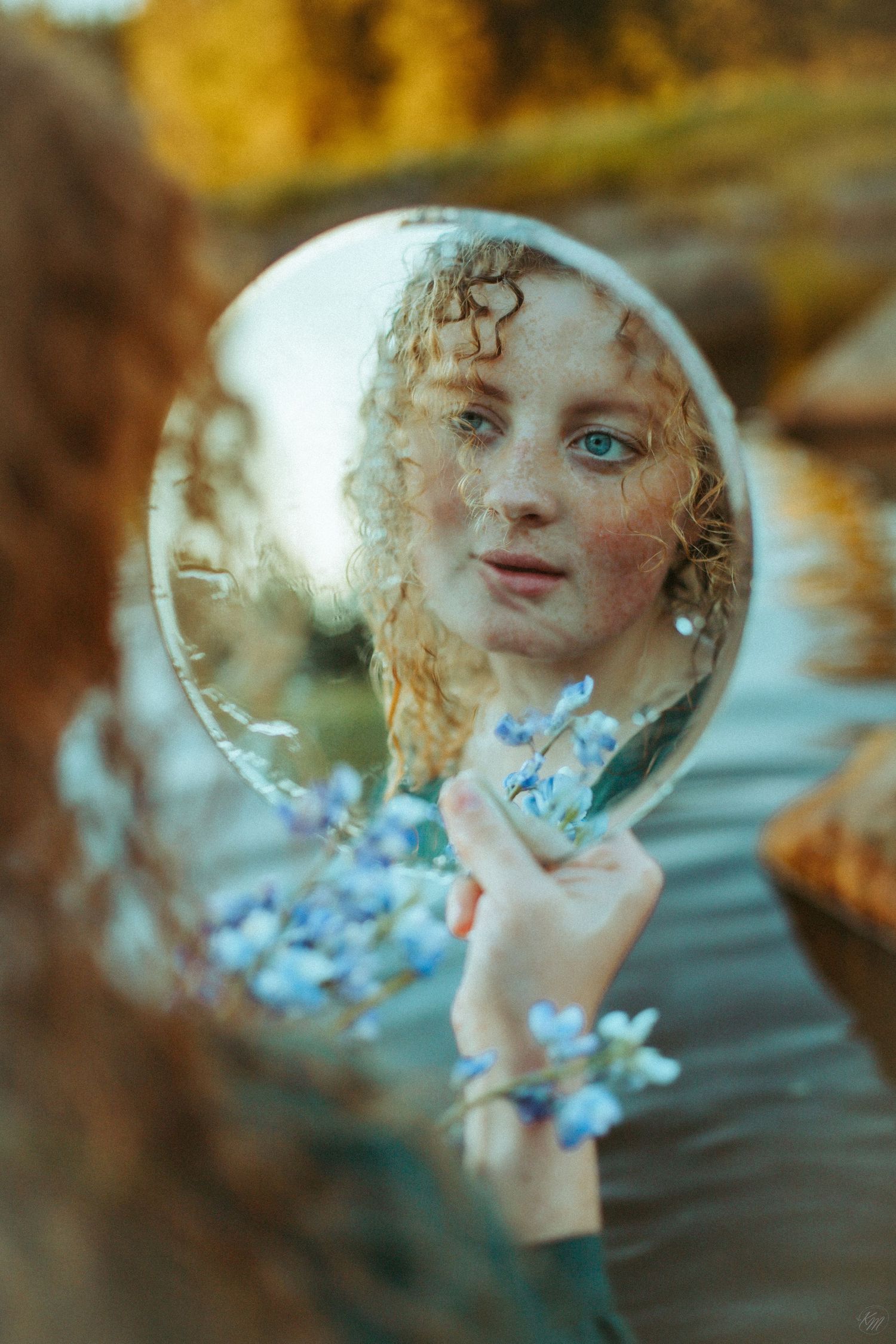 Reflection in circular mirror held near blue flowers by water's edge.