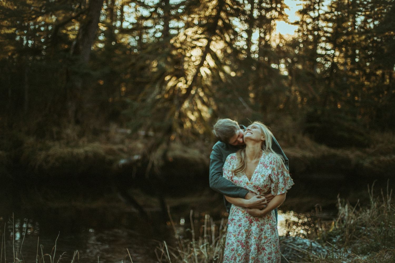 Couple in floral dresses embrace near a tranquil pond at sunset with evergreen forest in the background in Southeast Alaska