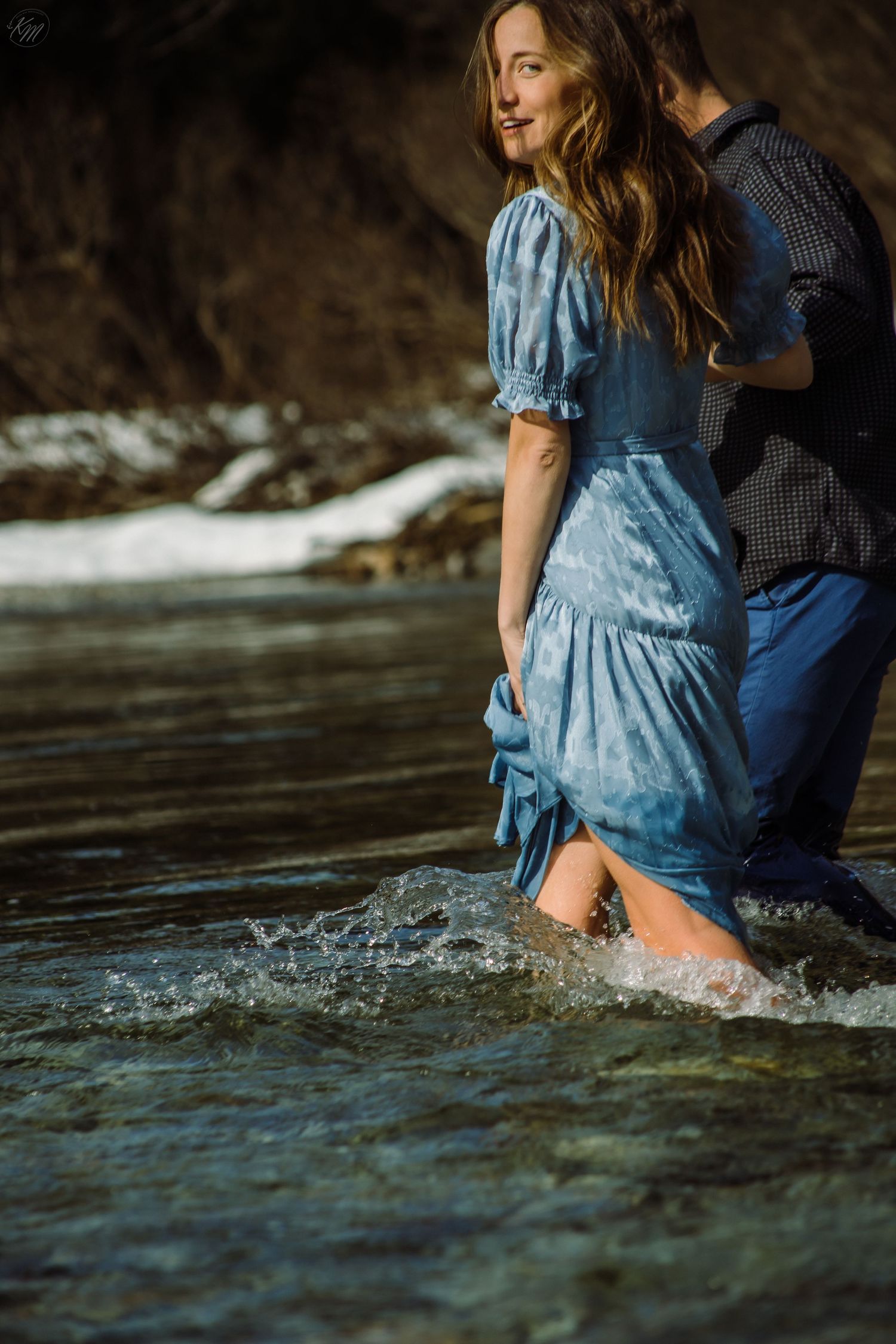 Couple wading in mountain river with woman wearing flowing blue dress.