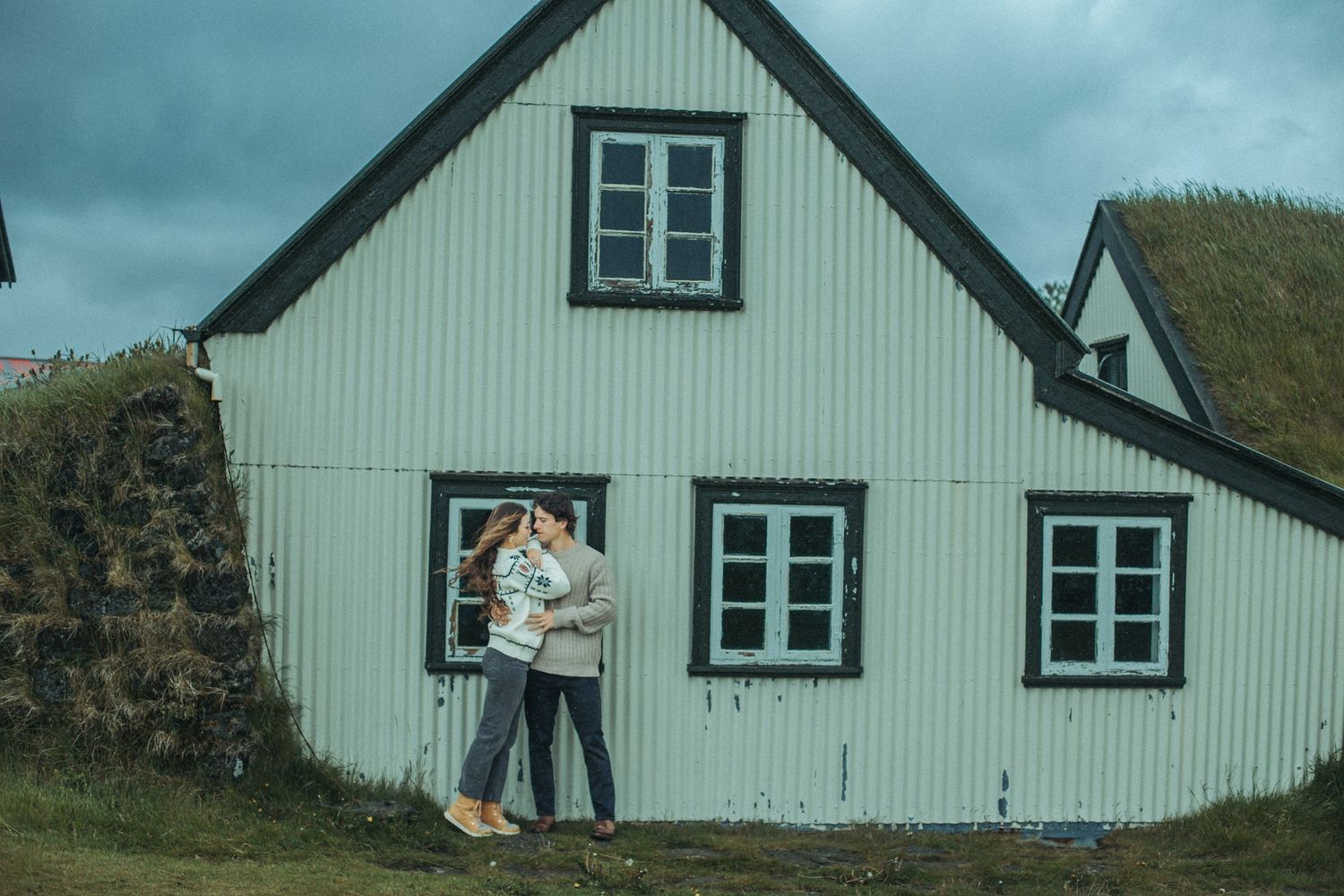 Couple in winter wear posing playfully by a white Nordic-style turf house while wearing cozy sweaters in Iceland for Engagement.
