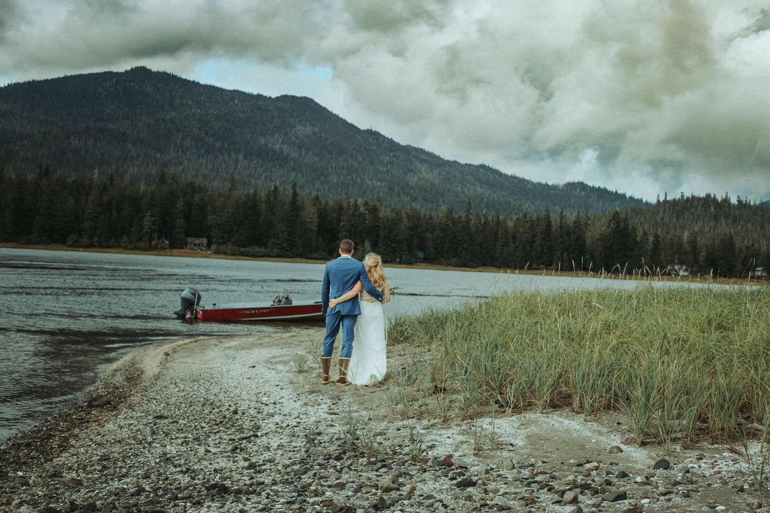 Bride and groom holding hands by ocean with red canoe and mountain views in Southeast Alaska.