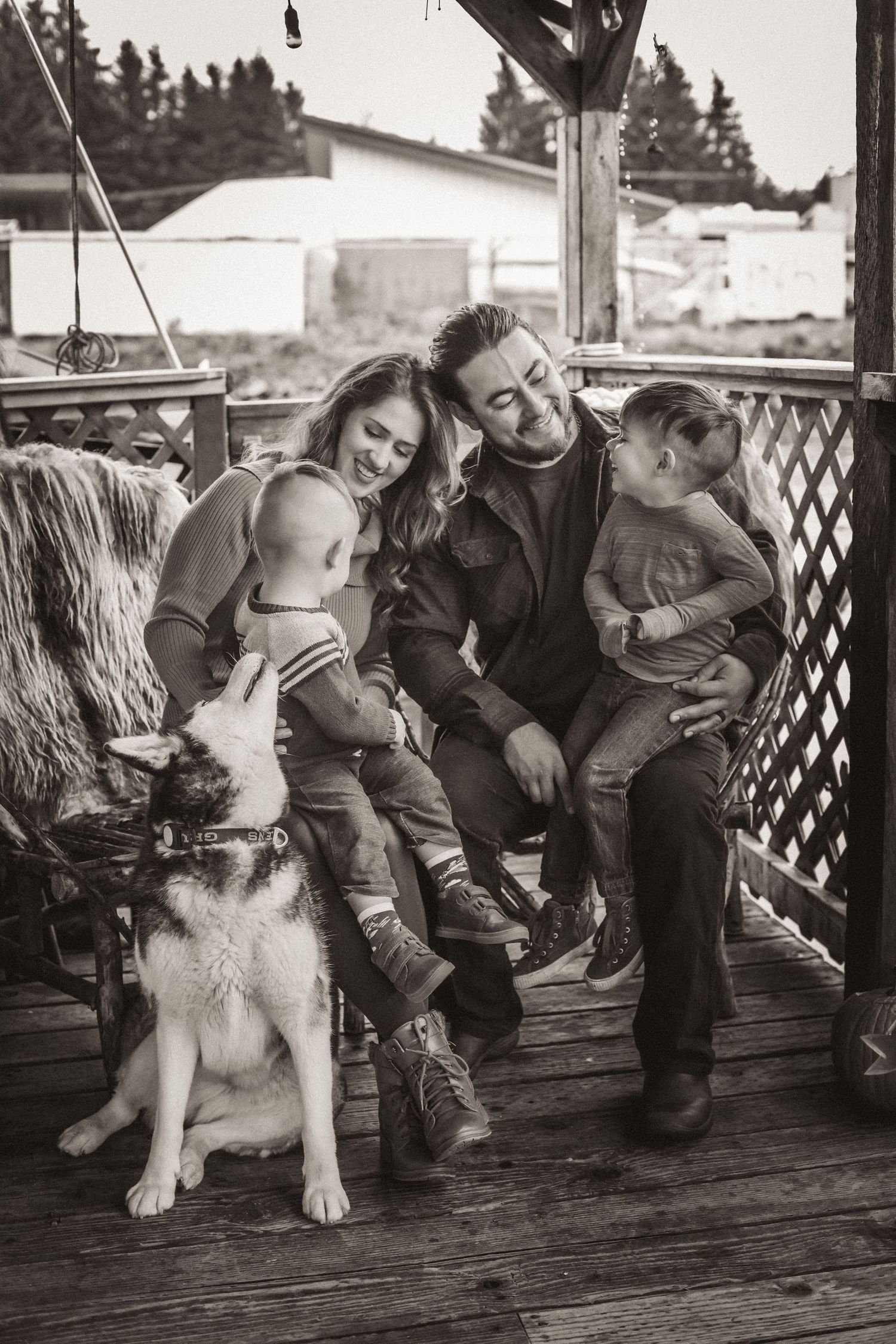 Black and white family portrait with two kids and husky dog on wooden porch.
