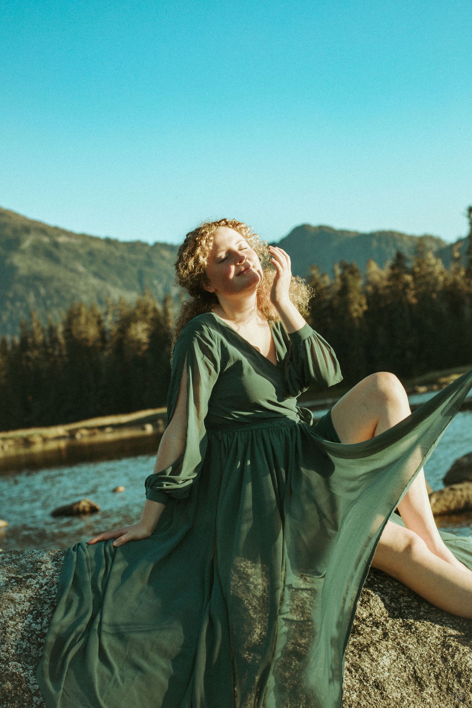 Woman in flowing green dress poses on rock by mountain lake during golden hour.