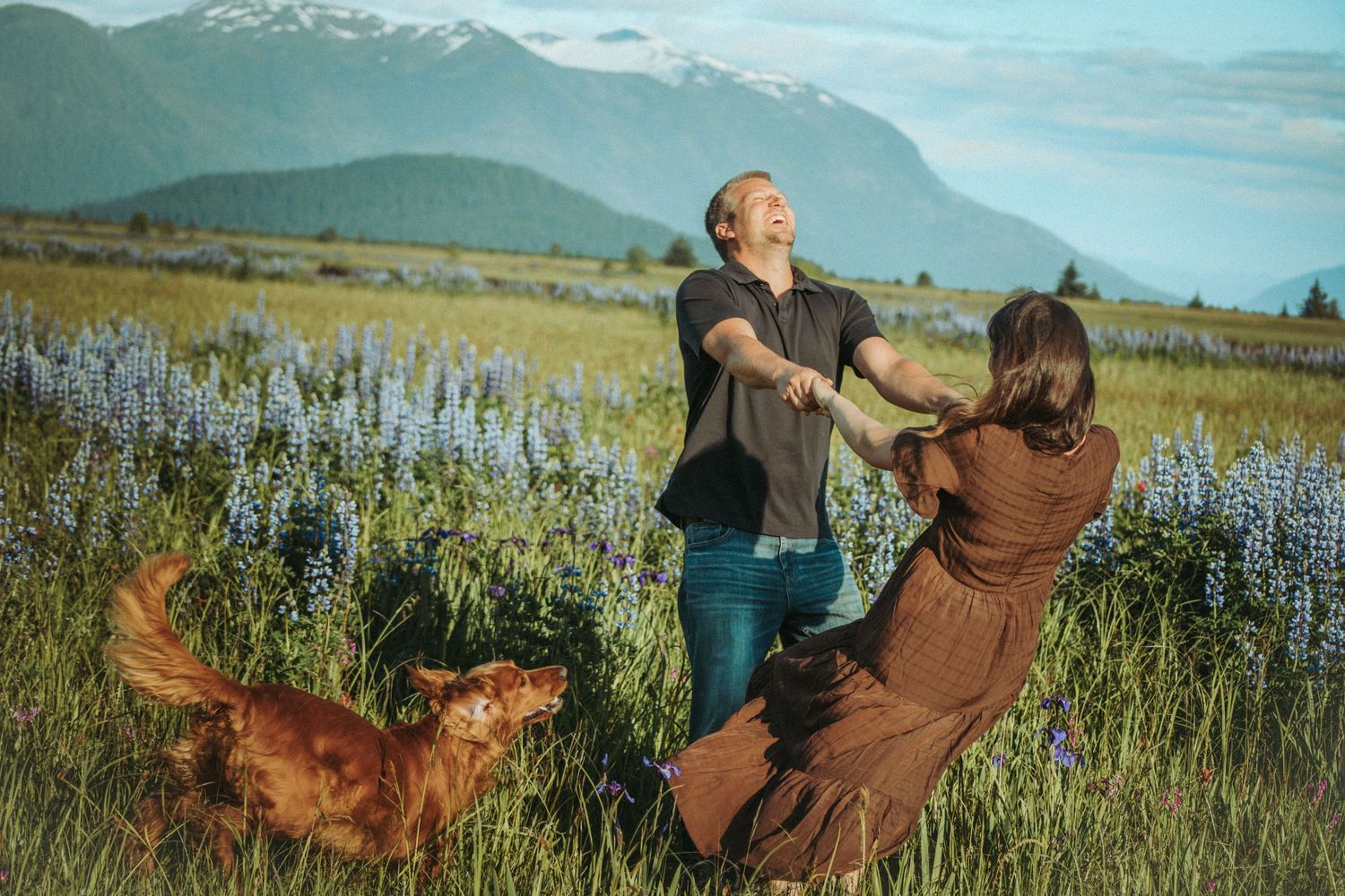 Engaged couple dance together in a wildflower meadow with mountains in the background as their dog plays nearby. SoutheastAlaska and lupine.