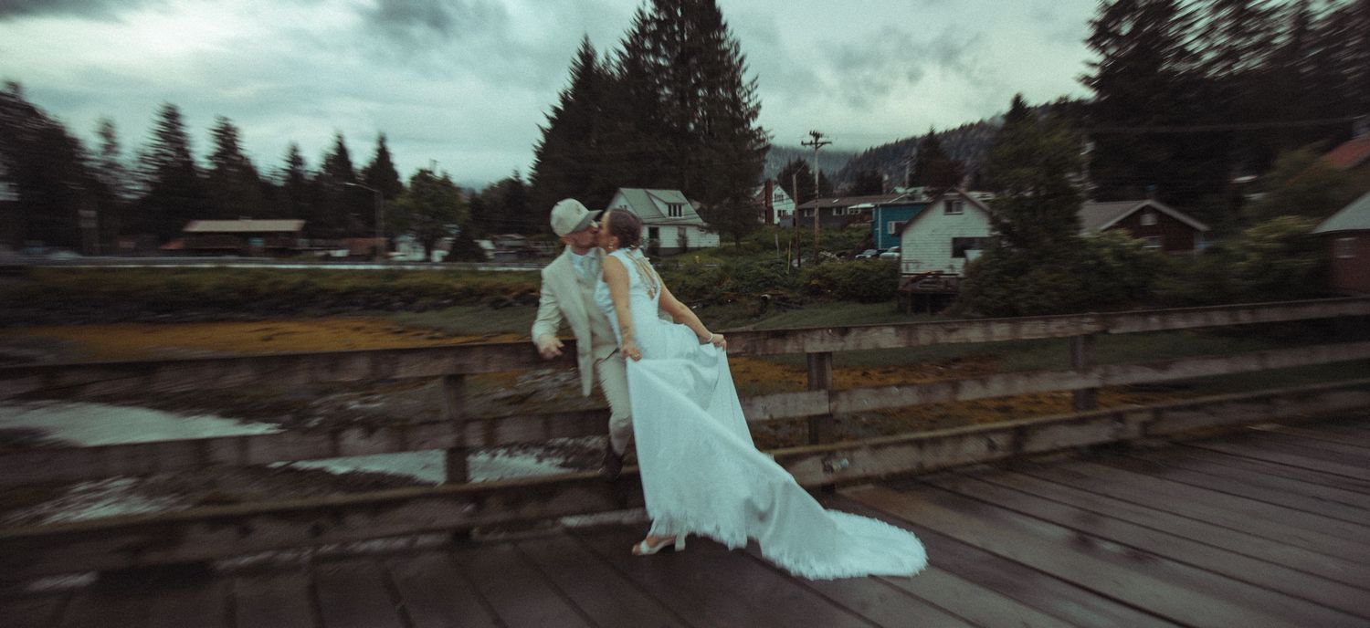 A bride and groom share a romantic moment on a wooden bridge in a rainy Pacific Northwest town setting.