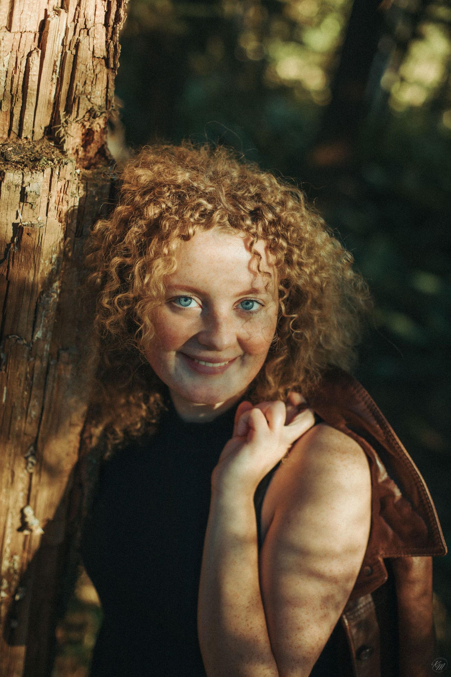 Portrait against a tree trunk wearing a black top and brown jacket in a forest setting.