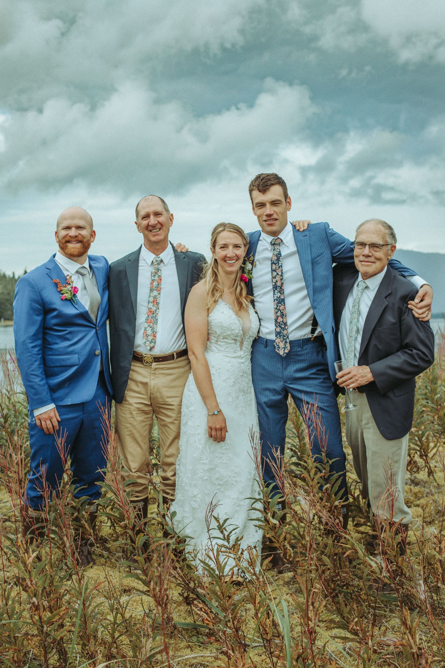 Small wedding party gathering for a formal photo in a field with mountains and cloudy sky.