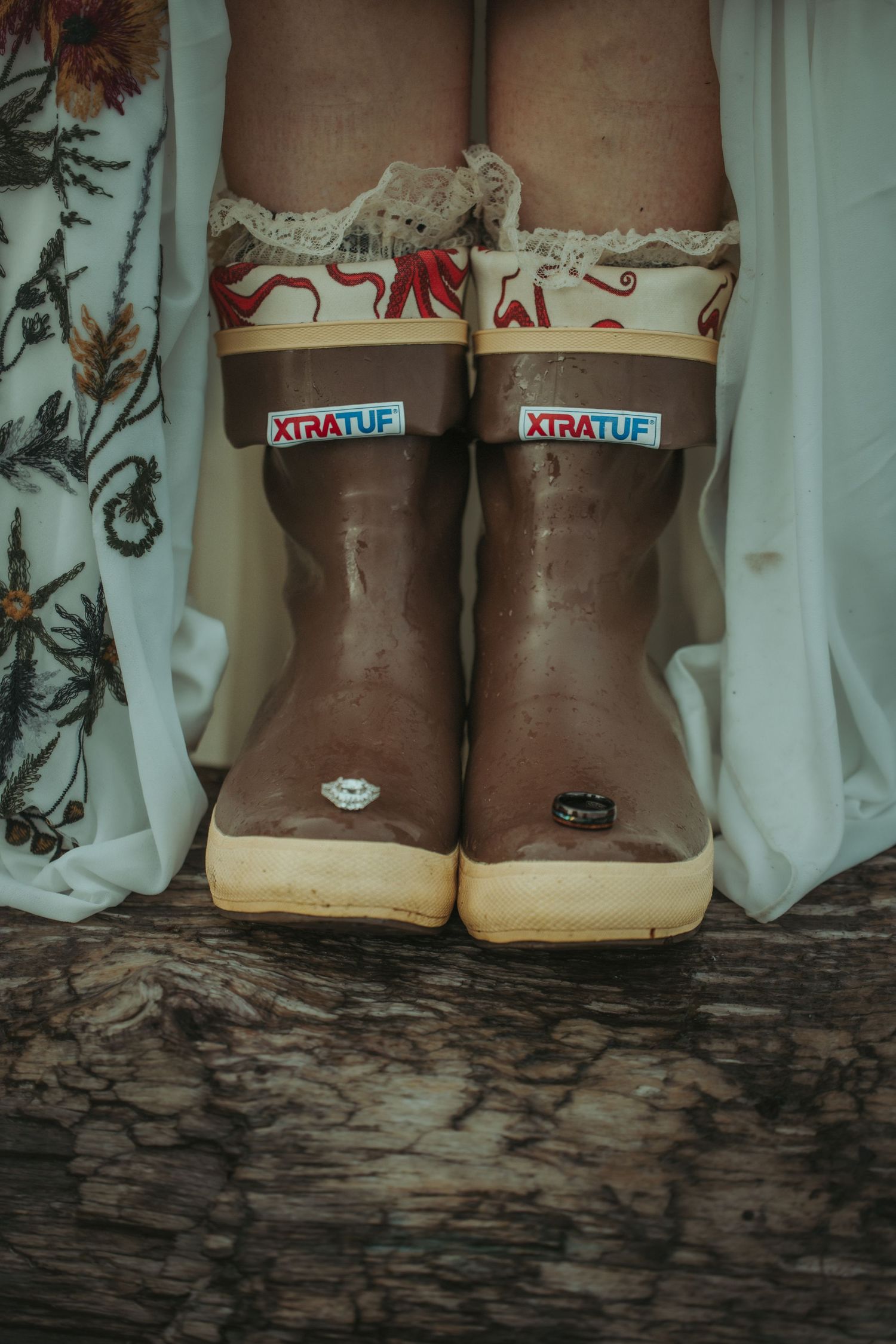 Bride wearing white lace dress with XtraTuf brown rubber boots standing on wooden surface.
