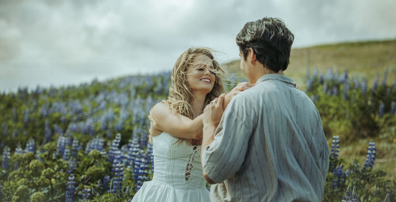 Blonde woman in white dress smiling at man in lupine flower field under cloudy sky.