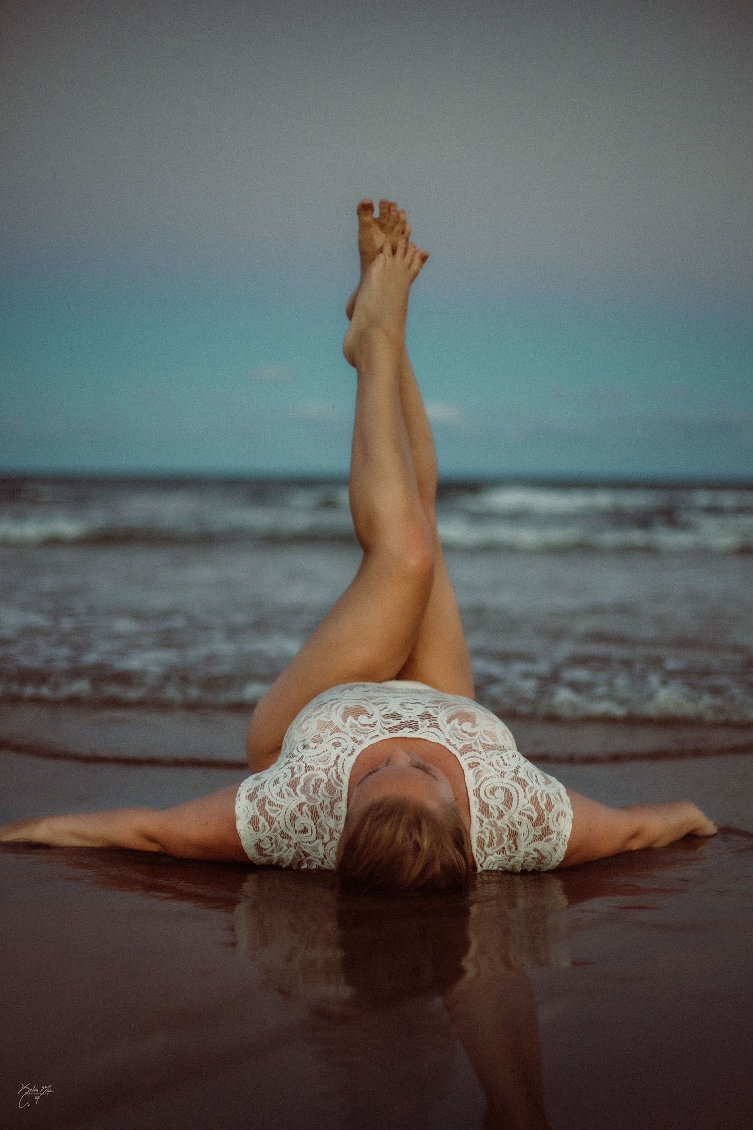 Woman in white lace bodysuit lying on wet beach with legs raised against ocean backdrop.