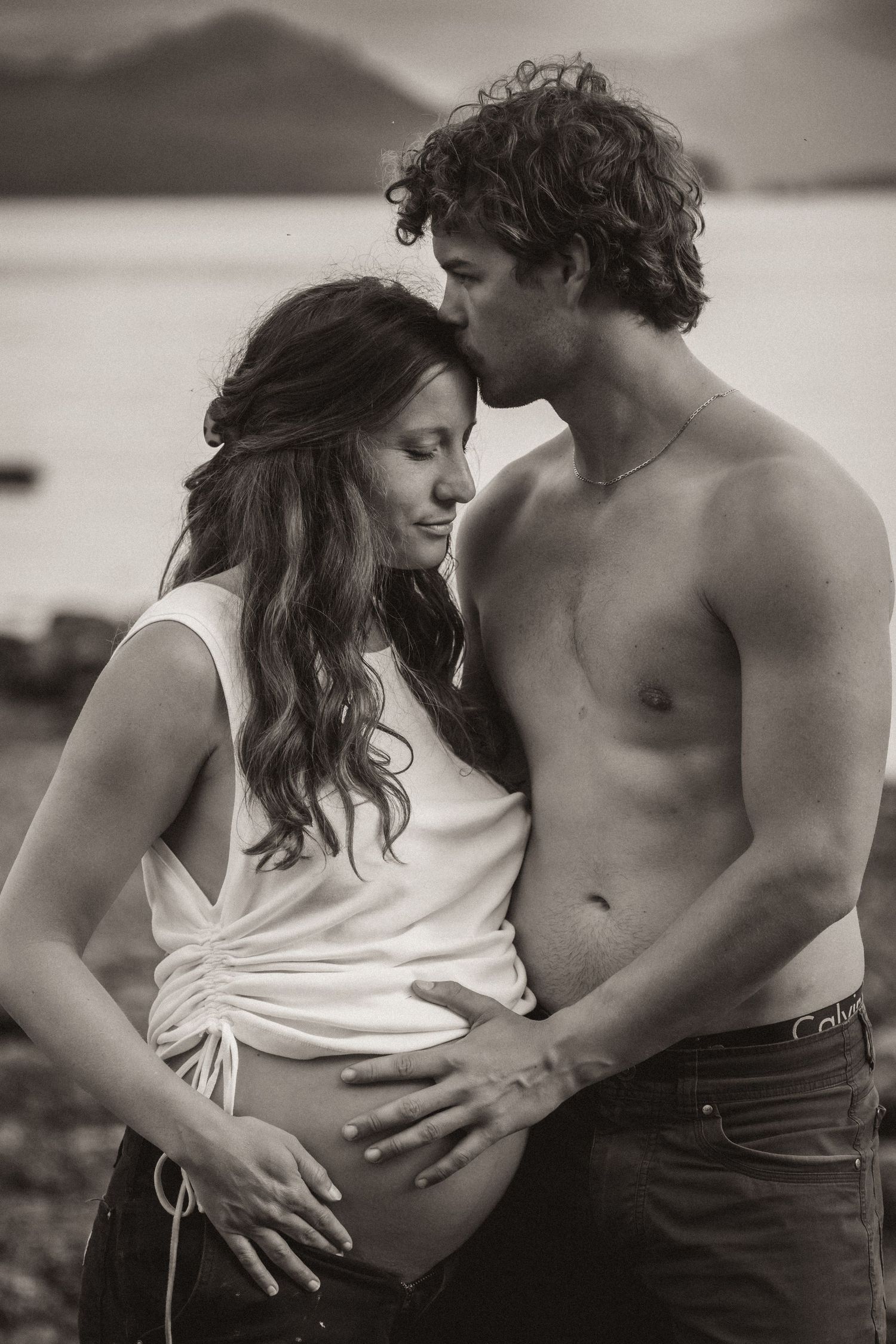 Black and white romantic maternity photo of couple embracing by mountain lake in Alaska.