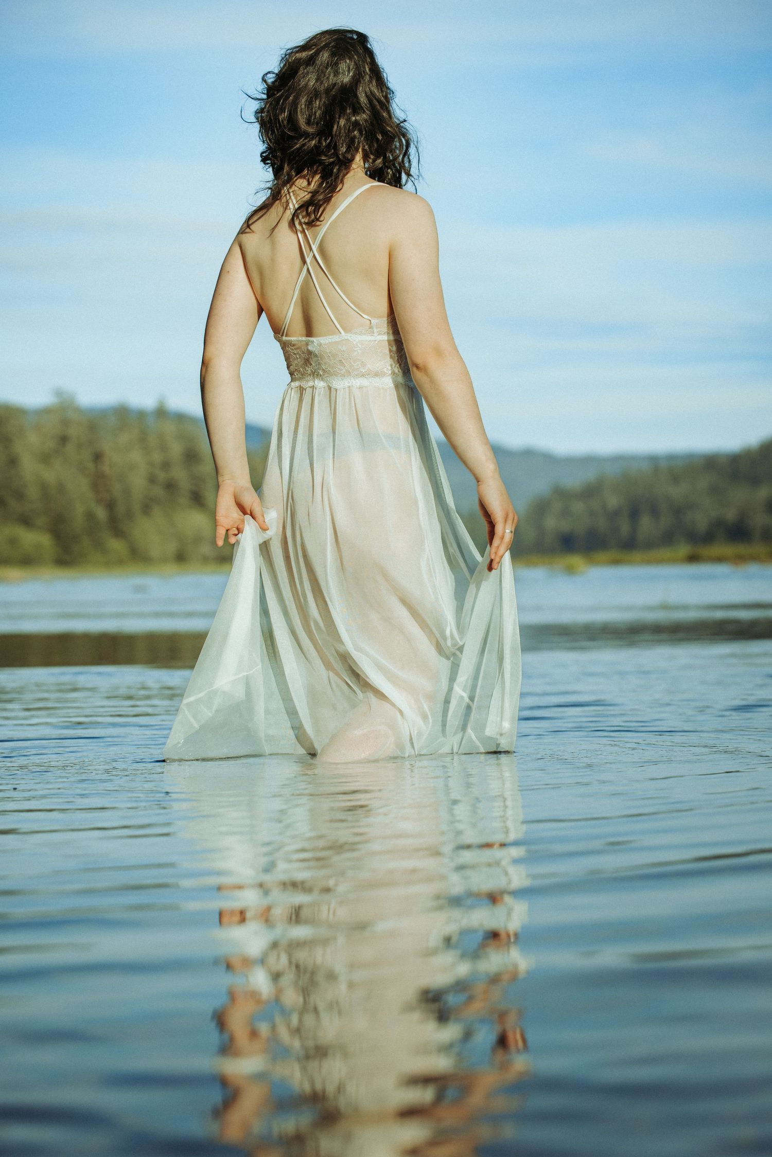 Woman in sheer white gown wading in a calm lake with forested hills in the distance.