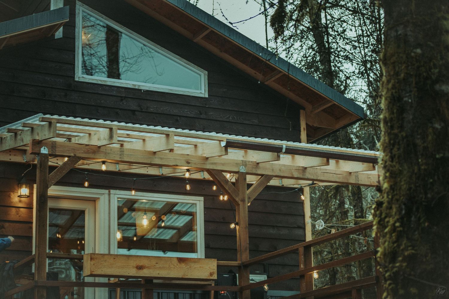 A rustic wooden cabin with a pergola porch and string lights at dusk creates a cozy outdoor atmosphere in Southeast Alaska.