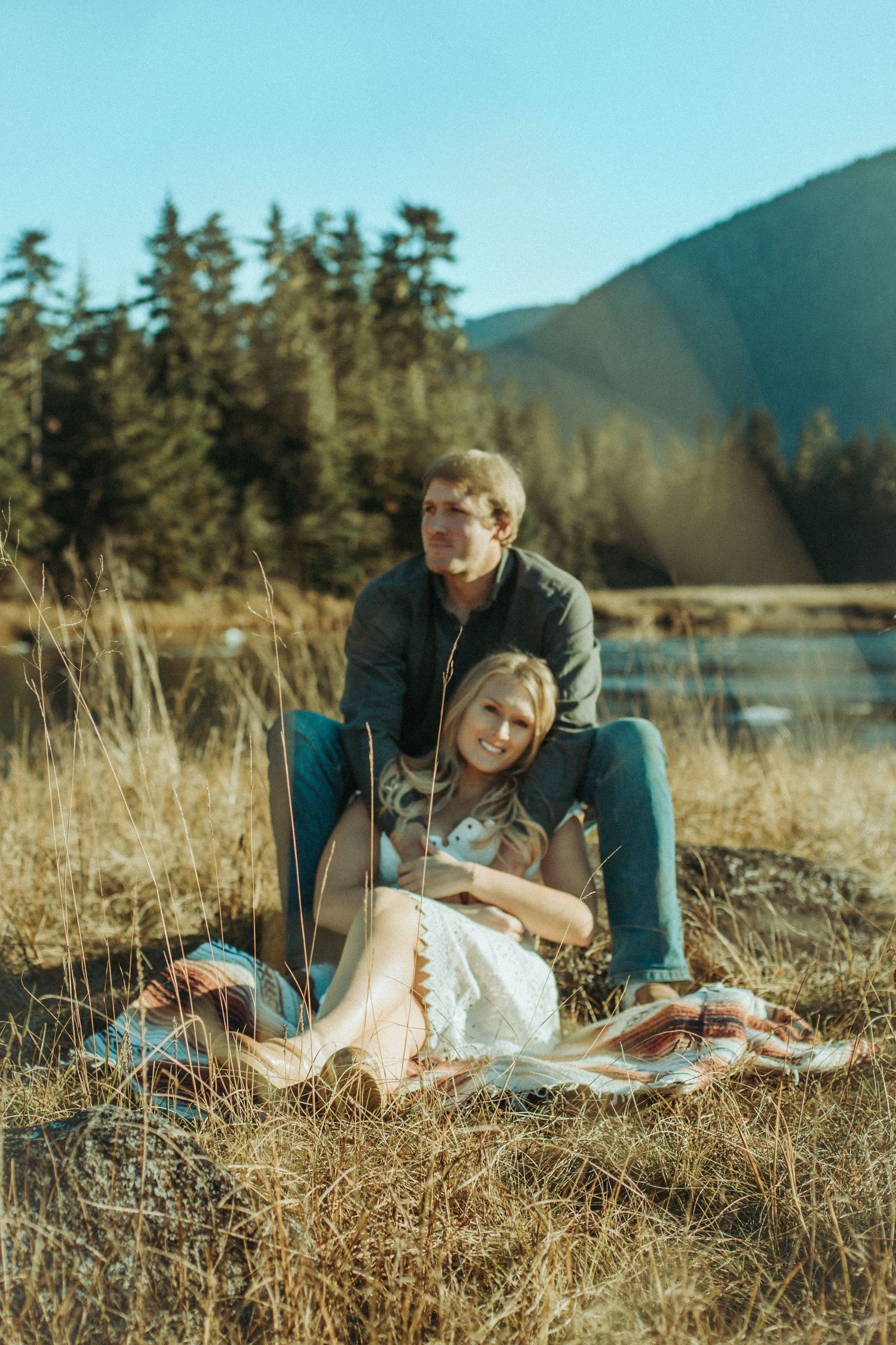 Couple sitting together in golden field with evergreen trees and mountains in background on sunny autumn day in Southeast Alaska.
