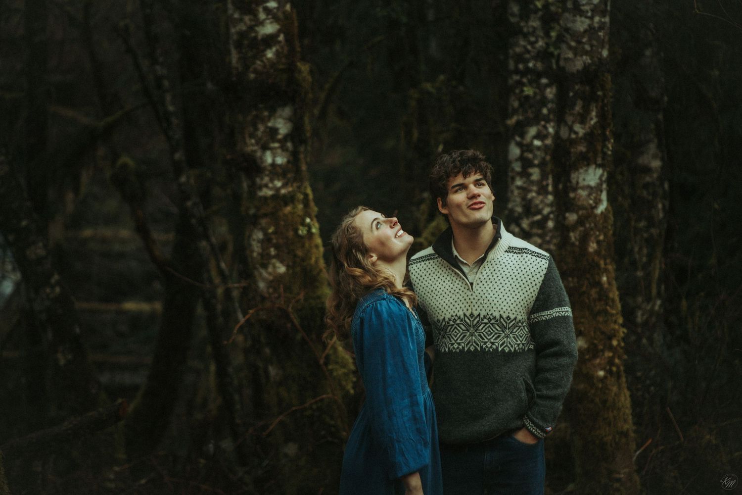 A loving couple stands close together in a moody forest with dark trees and muted lighting in Southeast Alaska.