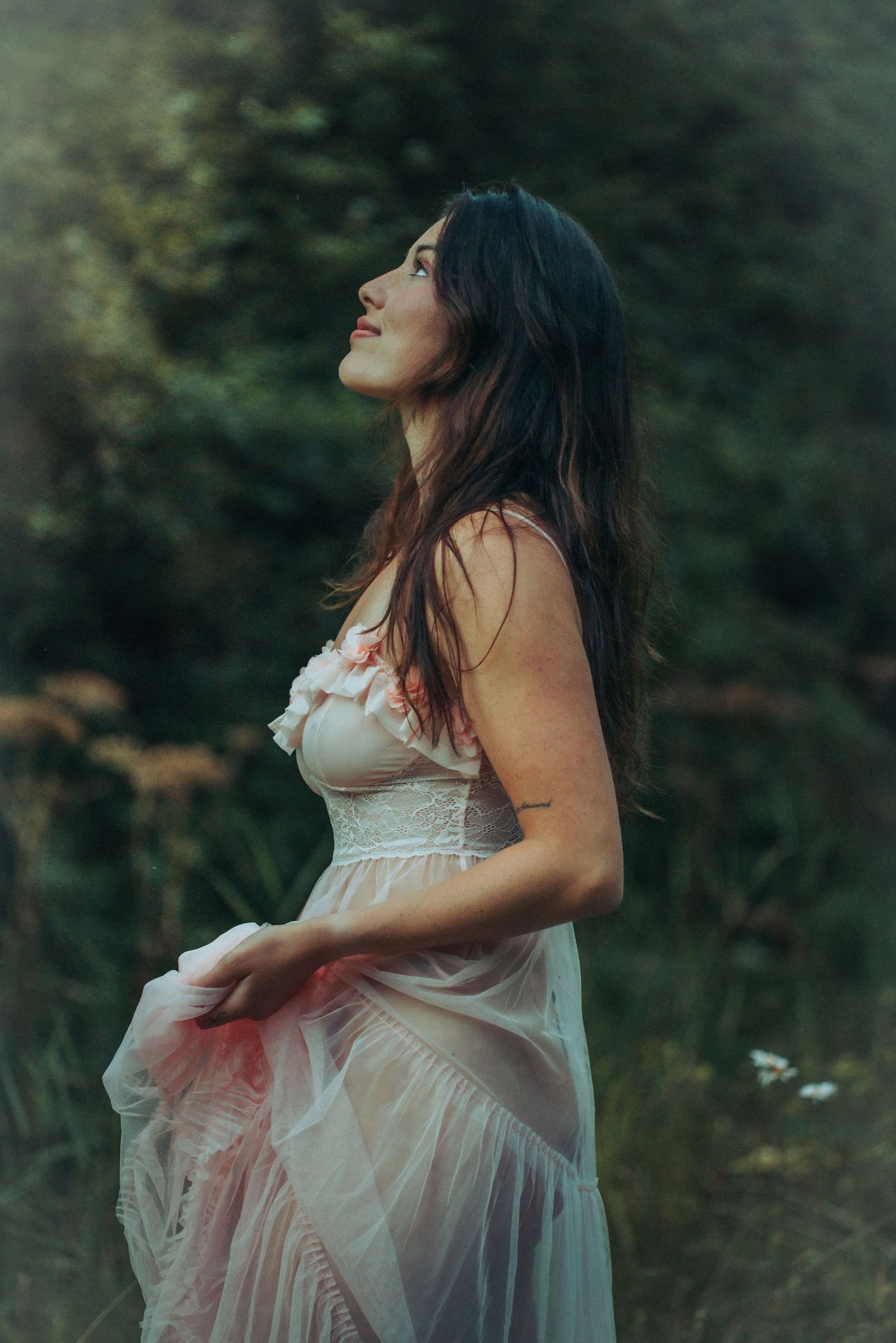 Dark-haired woman in sheer pink dress gazing upward in a lush green forest setting.