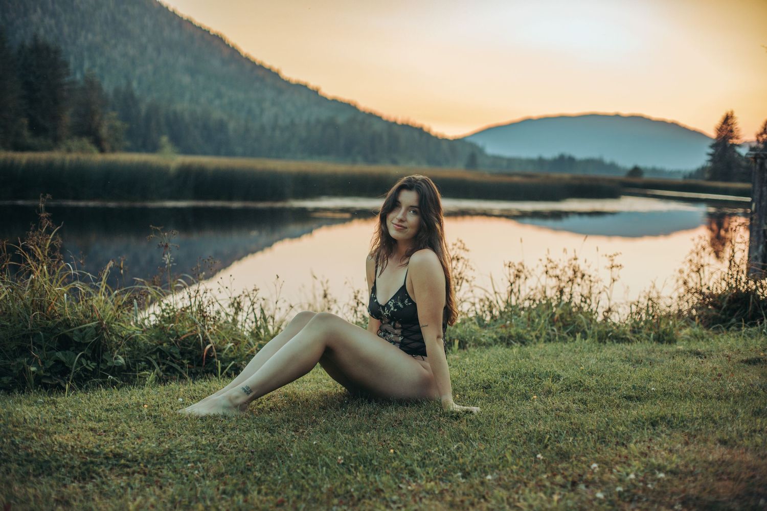 Woman in swimsuit sitting on grass by a serene mountain lake at sunset with forested hills in background.